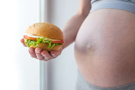 A pregnant woman with belly by the window holding a burger. The concept of fast food and unhealthy eating during pregnancyの写真素材