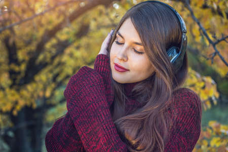 Beautiful woman in autumn Sunny Park standing listening to music with her headphones on the background of nature. The concept of good mood, favorite music, happy time.の写真素材