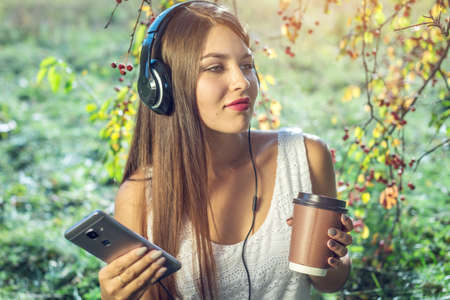 Attractive woman listening to music in your phone wearing headphones and holding a Cup of coffee in hand on a Sunny day in the Park. The concept of audiobooks and student educationの写真素材