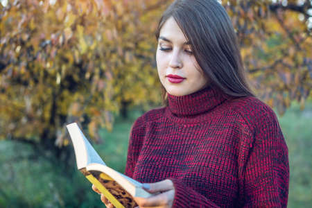Beautiful woman reading a novel holding a book in a Park on a background of yellow-orange trees. The concept of autumn romantic moodの写真素材