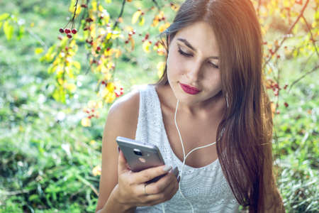 Attractive woman listening to music in your phone wearing headphones on a Sunny day in the Park. The concept of audiobooks and student educationの写真素材
