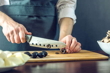 The chef in black apron cuts black olives with a knife. The concept of eco-friendly products for cookingの写真素材