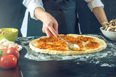The chef in black apron makes pizza with his hands putting the ingredients for the pizza on the table. The dark backgroundの写真素材