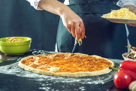 The chef in black apron makes pizza with his hands putting the ingredients for the pizza on the table. The dark backgroundの写真素材