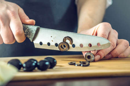 The chef in black apron cuts black olives with a knife. The concept of eco-friendly products for cookingの写真素材