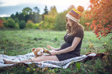 An expectant mother with a tummy sits on a blanket with a toy bear and reading stories to the baby in a beautiful autumn Park. The concept of pregnancy and the autumn harmonyの写真素材