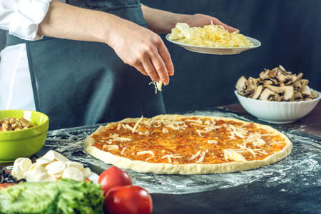 The chef in black apron makes pizza with his hands putting the ingredients for the pizza on the table. The dark backgroundの写真素材