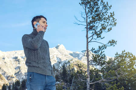 A young man in a sweater with phone in hand on the snowy mountain far from civilization on a background of blue sky. The concept of activity and the availability of mobile connectionの写真素材