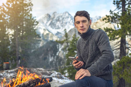 Man sits on the nature of the campfire in a sweater talking on the phone. Background mountain snowy peaks. Concept of mobile communication the Hiking and outdoor campingの写真素材