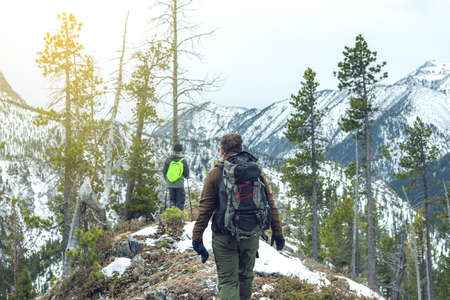 Men hikers with a backpack on top of the mountain back, looking at the snow slope. The concept of active relaxation, motivation and goal achievementの写真素材