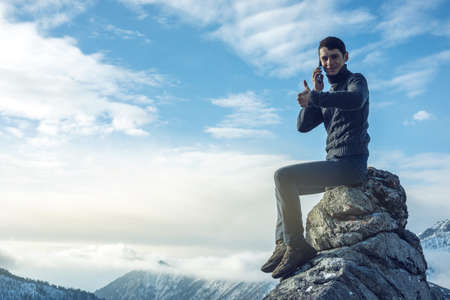 A young man in a sweater with phone in hand on the top of a snowy mountain far from civilization on a background of blue sky. The concept of activity and the availability of mobile connectionの写真素材