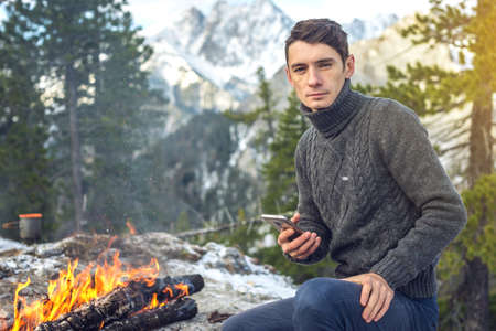 Man sits on the nature of the campfire in a sweater talking on the phone. Background mountain snowy peaks. Concept of mobile communication the Hiking and outdoor campingの写真素材