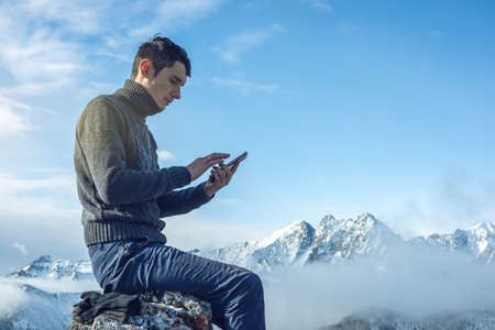 A young man in a sweater with phone in hand on the top of a snowy mountain far from civilization on a background of blue sky. The concept of activity and the availability of mobile connectionの写真素材
