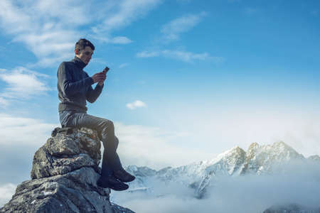 A young man in a sweater with phone in hand on the top of a snowy mountain far from civilization on a background of blue sky. The concept of activity and the availability of mobile connectionの写真素材