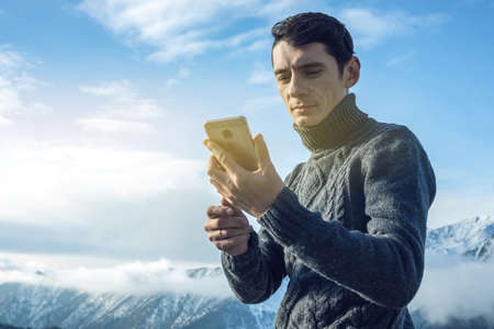 A young man in a sweater with phone in hand on the top of a snowy mountain far from civilization on a background of blue sky. The concept of activity and the availability of mobile connectionの写真素材