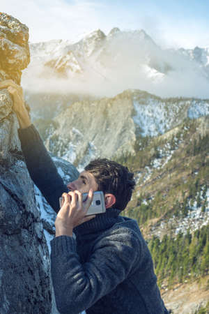 A young man climbs the mountain to the top and talking holding the phone in his hand. The concept of persistence and goal achievement in your career. Work in difficult conditionsの写真素材