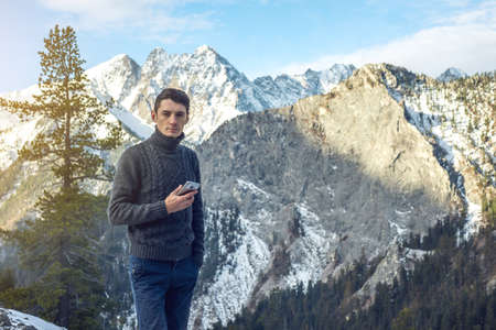 A young man in a sweater with phone in hand on the snowy mountain far from civilization on a background of blue sky. The concept of activity and the availability of mobile connectionの写真素材