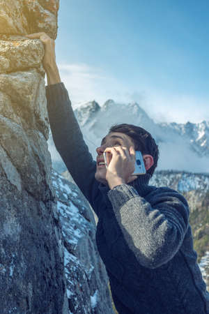 A young man climbs the mountain to the top and talking holding the phone in his hand. The concept of persistence and goal achievement in your career. Work in difficult conditionsの写真素材