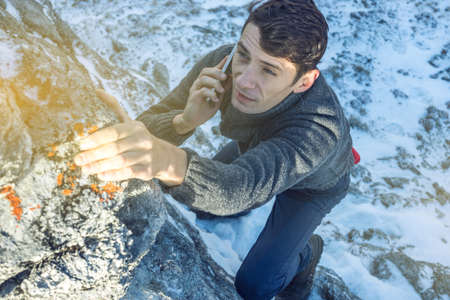 A young man climbs the mountain to the top and talking holding the phone in his hand. The concept of persistence and goal achievement in your career. Work in difficult conditionsの写真素材