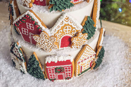 Large tiered Christmas cake decorated with gingerbread cookies and a house on top. Tree and garlands in the background. The concept of The desserts for the new yearの写真素材