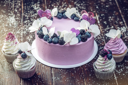 Purple beautiful cake decorated with berries, blackberries and blueberries on top with cupcakes on...