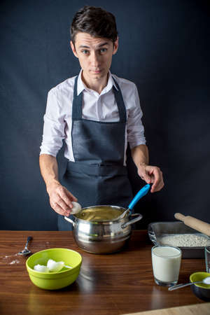 Young man chef in uniform black apron cooking cake with ingredients on the table in the kitchen. Recipes by stepsの写真素材