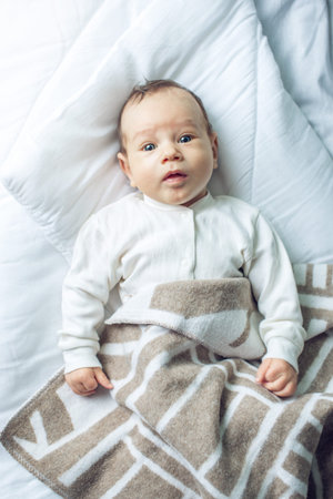 Cute funny baby lying on a white bed covered with a blanket. The concept of The tenderness of...
