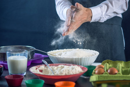 Chef pastry in a black apron in the process of making cupcakes from ingredients on dark background....