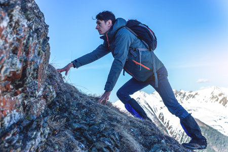 Man Hiking with a backpack crawling on rocky rocks to the top on the background of snowy mountains. Concept the hard way and goal achievementの写真素材