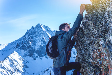 Man Hiking with a backpack crawling on rocky rocks to the top on the background of snowy mountains. Concept the hard way and goal achievementの写真素材