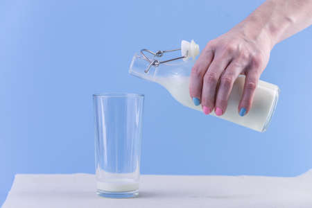 A woman's hand pours fresh milk from a glass bottle into a glass on a blue background.の写真素材