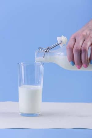 A woman's hand pours fresh milk from a glass bottle into a glass on a blue background.の写真素材