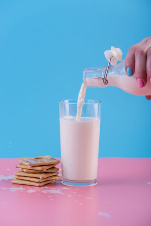 A woman's hand pours fresh milk from a glass bottle into a glass and cookies on a pastel background.