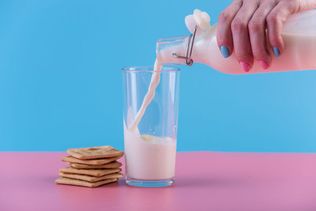 A woman's hand pours fresh milk from a glass bottle into a glass and cookies on a pastel background.