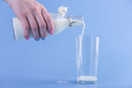 A woman's hand pours fresh milk from a glass bottle into a glass on a blue background.の写真素材