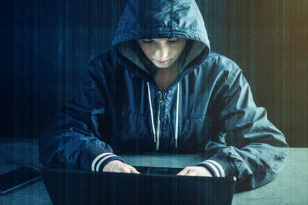 Hacker teen programmer uses a laptop to hack the system. Stealing personal data. Creation and infection of malicious virus. The concept of cyber crime and hacking electronic devicesの写真素材
