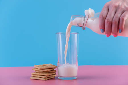A woman's hand pours fresh milk from a glass bottle into a glass and cookies on a pastel background. Colorful minimalism. The concept of healthy dairy products with calciumの写真素材