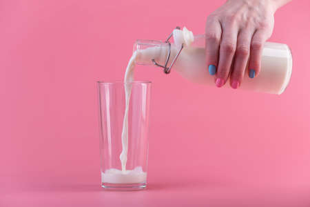 A woman's hand pours fresh milk from a glass bottle into a glass on a pink background. Colorful minimalism. The concept of healthy dairy products with calciumの写真素材