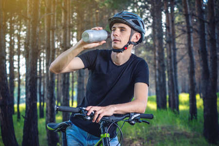 A cyclist in a helmet drinking water from a bottle while riding in the Park among the trees. Concept of active and healthy lifestyleの写真素材