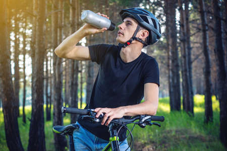 A cyclist in a helmet drinking water from a bottle while riding in the Park among the trees. Concept of active and healthy lifestyleの写真素材
