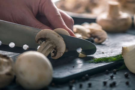 Women's hands are cutting champignons with a knife on the dark kitchen table. The concept of cooking mushroom dishes. Close upの写真素材