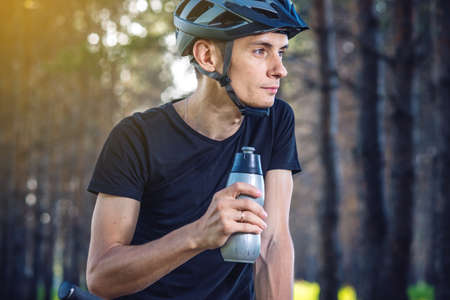 A cyclist in a helmet drinking water from a bottle while riding in the Park among the trees. Concept of active and healthy lifestyleの写真素材