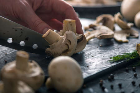 Women's hands are cutting champignons with a knife on the dark kitchen table. The concept of cooking mushroom dishes. Close upの写真素材