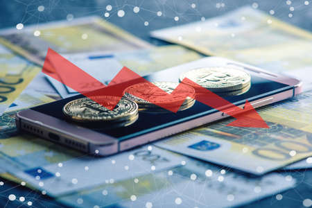 Physical bitcoin coin on the phone screen on the background of the Euro banknotes. Red arrow of the chart of falling quotes downの写真素材