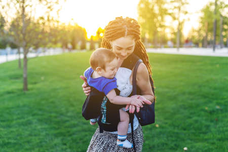 Young happy mom with baby son in ergo backpack walking in the Park. Sunny summer day. The concept of modern parentsの写真素材