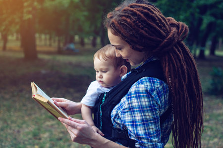 Beautiful mom with a baby in ergo backpack reading a fairy tale in the Park. Children's education in...