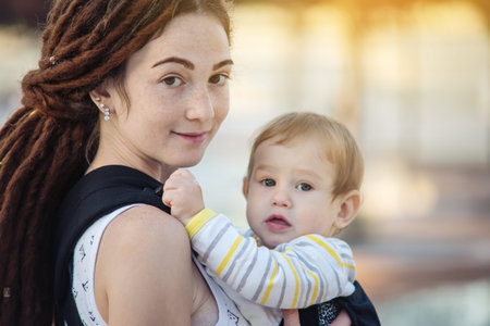 Young modern happy mother with baby son in ergo backpack walking in Sunny summer day. The concept of...