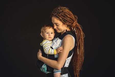 Portrait young happy mom with baby son in ergo backpack on dark wall on the background. The concept of modern parentsの写真素材