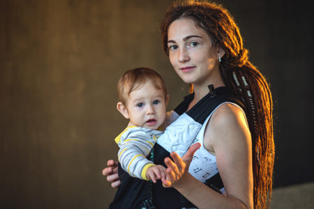 Portrait young happy mom with baby son in ergo backpack on dark wall on the background. The concept...