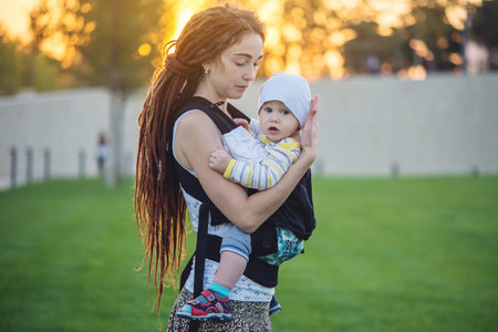 Young modern happy mom with baby son in ergo backpack walking in Sunny summer day. The concept of...